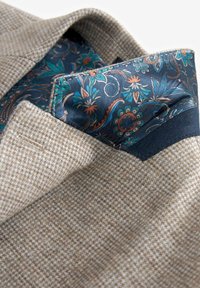 Beige gemusterter Stoff mit einer strukturierten Oberfläche. Der Kragen innen zeigt ein marineblaues Blumenmuster mit Akzentfarben in Blau, Orange und Grün.