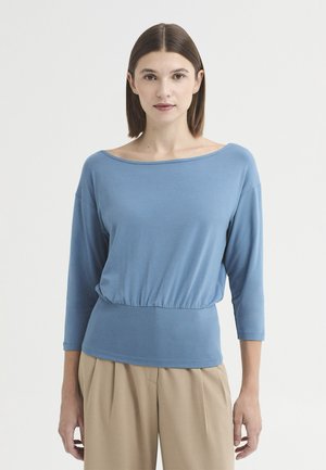 Femme aux cheveux bruns raides portant un haut bleu à manches longues et un pantalon beige, debout devant un fond clair uni.