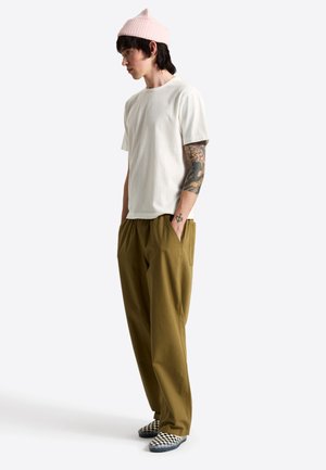 Ung mand iført en pink beanie, hvid t-shirt, olivengrønne bukser, ternede slip-on sko og en farverig tatovering på venstre arm.