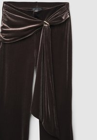 Pantaloni in velluto marrone scuro con fronte drappeggiato, caratterizzati da un dettaglio in metallo color oro al centro. Texture liscia, design a vita alta.