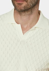 Krämfärgad stickad polo med ett diamantmönster, med en klassisk krage och strukturerad tyg. Det finns inslag av subtil glans.