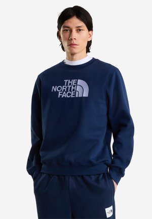 Junger Mann trägt ein marineblaues The North Face Sweatshirt und passende Hose, steht mit den Händen in den Taschen vor weißem Hintergrund.