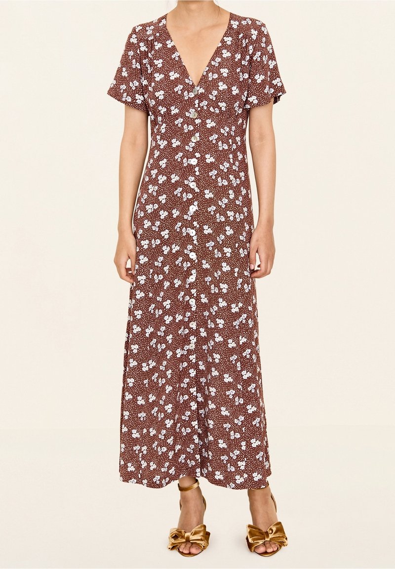 Femme portant une robe midi marron à fleurs boutonnée avec manches courtes et des talons ouverts dorés avec de grands nœuds.