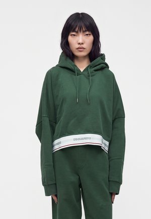 Jonge vrouw met steil zwart haar die een groene cropped hoodie en bijpassende broek draagt tegen een effen lichte achtergrond.
