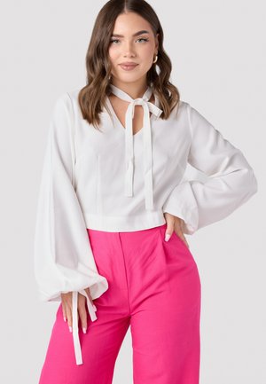 White blouse with a V-neck and front tie detailing, featuring long balloon sleeves. Paired with vibrant pink high-waisted trousers.
