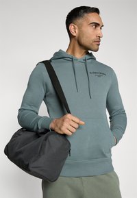 Felpa con cappuccio in teal smorzato, caratterizzata da una tasca a marsupio e un logo. Borsone sportivo nero con superficie texturizzata tenuto in mano.