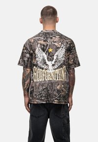T-shirt w kamuflażowym wzorze w stonowanych kolorach, z białymi grafikami skrzydeł i napisem "GOODFORNOTHING" w żółtym kolorze na plecach. Krótkie rękawy, luźny krój.
