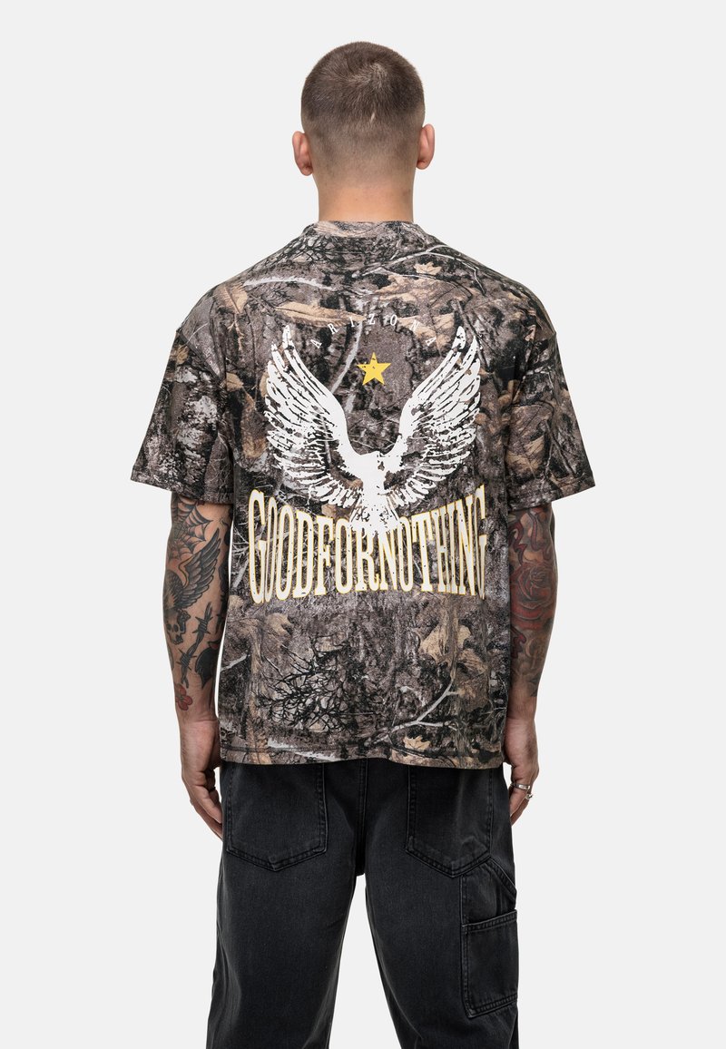 T-shirt w kamuflażowym wzorze w stonowanych kolorach, z białymi grafikami skrzydeł i napisem "GOODFORNOTHING" w żółtym kolorze na plecach. Krótkie rękawy, luźny krój.