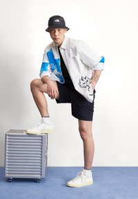 Bjela košulja s uzorkom i plavim detaljima, crne kratke hlače i bijele visoke tenisice. Model pozira na srebrnoj metalnoj kutiji protiv bijele pozadine.