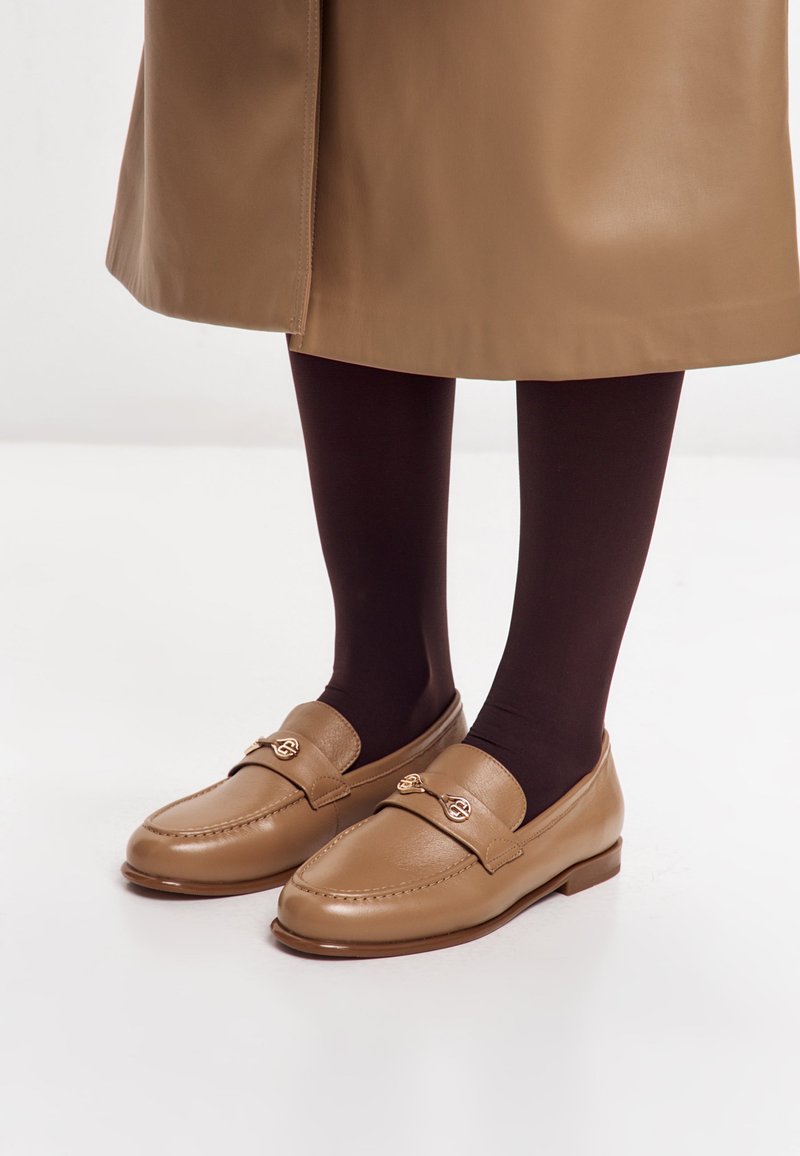 Hnědé kožené mokasíny s hladkým povrchem, zdobené zlatou kovovou ozdobou a plochou, zaoblenou špičkou.