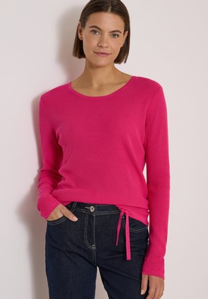Femme aux cheveux courts et bruns portant un haut à manches longues rose vif avec un nœud sur le côté et un jean bleu foncé, debout contre un mur clair.