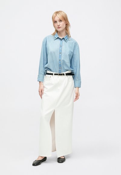 Donna che indossa una camicia di denim ricamata azzurro chiaro infilata in una lunga gonna bianca con spacco frontale, cintura nera e ballerine nere.