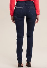Jean skinny en denim foncé avec une taille haute, comportant deux poches arrière et des détails de couture subtils, associés à des talons noirs.