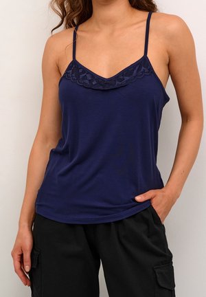 Vrouw in een marineblauw hemdtop met kantafwerking en zwarte cargobroek, staand met een hand in de zak tegen een effen achtergrond.