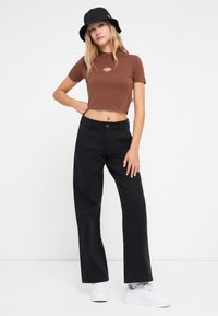 Brun cropped t-skjorte med logo, svarte vide bukser og hvite sneakers. Modellen har på seg en svart bøttehatt og står foran en ensfarget bakgrunn.