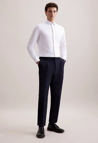 Camicia bianca a maniche lunghe con colletto abbottonato abbinata a pantaloni navy plissettati e scarpe nere, caratterizzata da una vestibilità su misura e un aspetto curato.