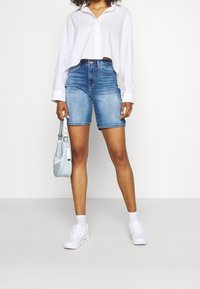 Camisa blanca de botones recortada con mangas largas, bermudas de denim azul, zapatillas blancas y un bolso ligero azul.