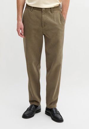 Mann trägt olivgrüne gerade geschnittene Hose mit elastischem Bund und schwarze Lederloafers, steht mit einer Hand in der Tasche.