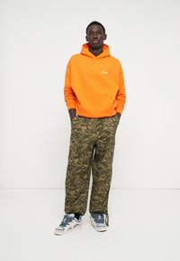 Felpa arancione brillante con tasca a marsupio, abbinata a pantaloni cargo con motivo mimetico verde e marrone, e sneakers azzurri chiari con accentuazioni scure.