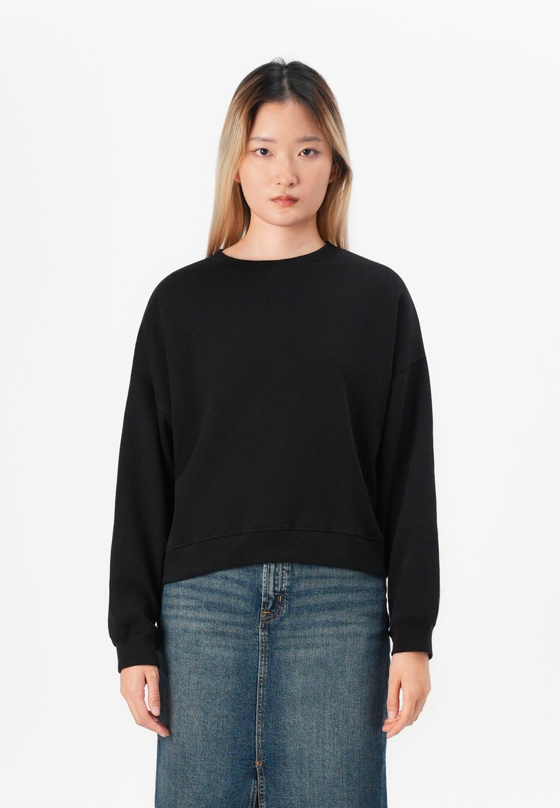 Sweatshirt noir court avec des manches longues, un col rond et des poignets côtelés, associé à une jupe en denim avec une fente à l'avant.