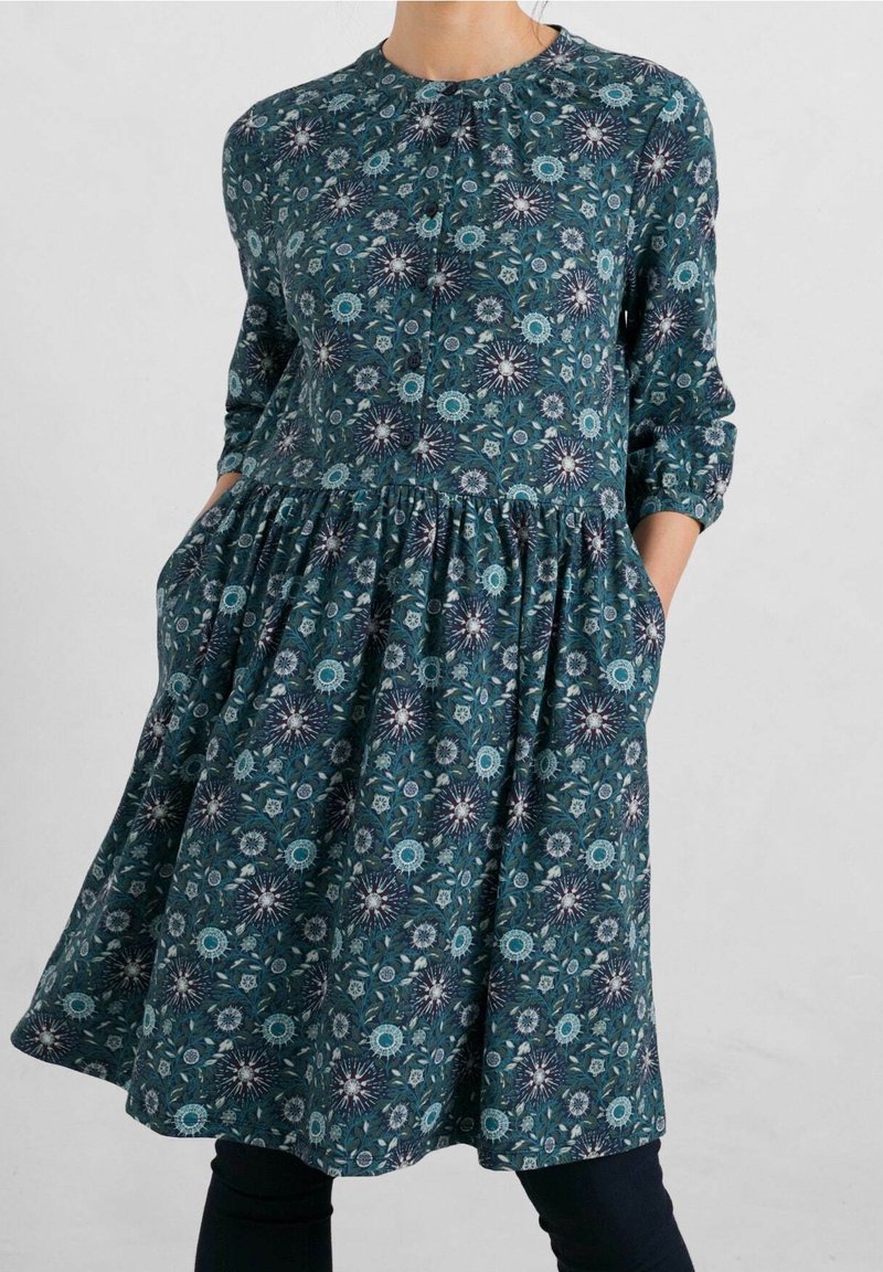 Robe bleu sarcelle à manches longues et taille froncée, avec un motif floral bleu clair et blanc, portée les mains dans les poches.