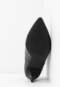 Chaussure noire à talons hauts et bout pointu avec motif texturé, semelle tournée vers l'avant, positionnée dans un angle contre un fond blanc.