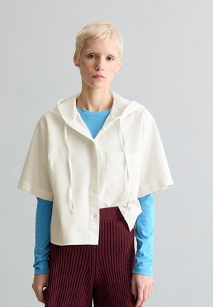 Person with short blond hair wearing a loose white short-sleeve hooded shirt over a blue long-sleeve top and maroon ribbed pants.