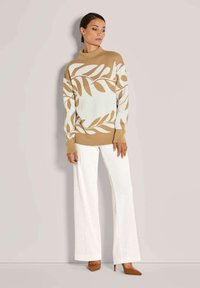 Frau, die einen beigen und weißen Pullover mit Blattmuster, weiße weit geschnittene Hose und braune spitz zulaufende High Heels trägt, steht vor einer schlichten Wand.