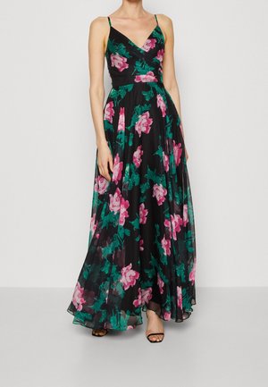 Kvinde iført en sort, ærmeløs maxikjole med pink og grøn blomsterprint og tynde skulderstropper, kombineret med sorte åbne hæle.