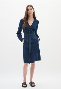 Denim omslagsklänning i mörkblått, med lång ärm och bälte. Har en V-ringning och två sidfickor. Slät textur och sömnadsdetaljer.
