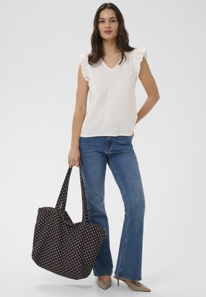 Žena v bielom volánovom tielku bez rukávov, modrých rifliach a béžových lodičkách, drží čiernu kabelku s bielymi bodkami, stojaca na bielom pozadí.