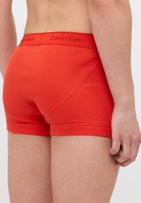 Crvene pamučne bokserice s mekim elastičnim pojasom na kojem se nalazi logo "Calvin Klein" u tamnocrvenoj boji. Glatka tekstura s prilagođenim dizajnom.