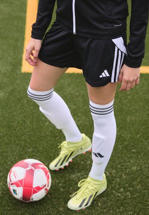 Schwarze Sportjacke, schwarze Shorts mit weißen Streifen, weiße Kniesocken und leuchtend gelbe Fußballschuhe; ein roter und weißer Fußball in der Nähe.