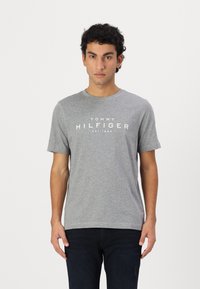 Grå kortermet T-skjorte laget av bomull, med hvit trykt tekst "TOMMY HILFIGER EST. 1985" på brystet.