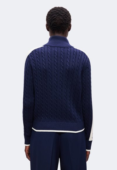 Personne portant un pull en maille torsadée bleu marine avec des accents blancs, vue de dos sur un fond clair uni.