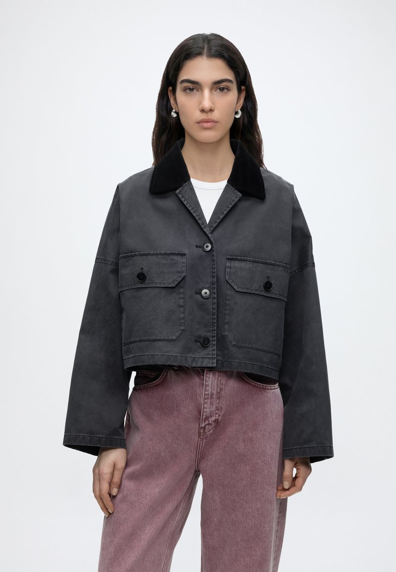 Femme aux longs cheveux foncés portant une veste courte noire boutonnée avec de grandes poches devant et un jean mauve délavé taille haute, debout sur un fond blanc.