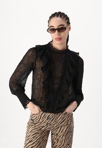 Zwarte doorzichtige blouse met gerimpelde randen en een stippenpatroon, voorzien van een neklijn en lange mouwen. Gecombineerd met panterprint broeken.