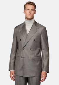 Double-breasted grey blazer with textured fabric, brown buttons, and patch pockets. Worn over a light grey turtleneck.