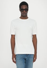 Hvit kortermet t-skjorte med rund hals og lett ribbet tekstur. Har en liten logo på venstre bryst. Kombinert med blå jeans.