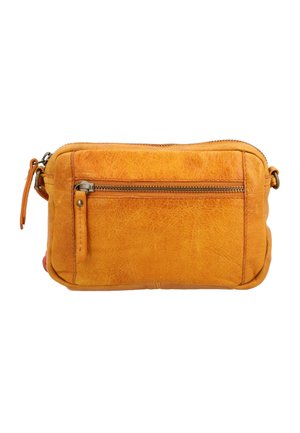 Uśmiechnięta kobieta w białej koszulce i niebieskich spodniach trzyma małą torebkę na ramię w kolorze musztardowej żółci z detalem na zamek.