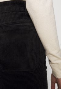 Tissu en velours côtelé noir avec des rayures verticales, montrant un détail de poche arrière et une partie d'un pull crème. Texture douce, design ajusté.