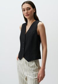 Gilet en lin noir avec un décolleté en V profond, comportant six boutons, une coupe ajustée et des coutures subtiles. Associé à un pantalon à rayures blanches.