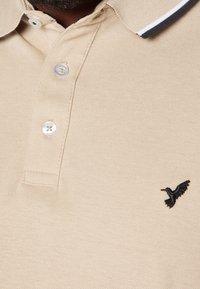 Beżowa koszulka polo o teksturowanym wykończeniu, z trzema guzikami i czarnym wyszywanym logo ptaka. Posiada kontrastowy, ciemny obwód kołnierzyka.