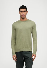 Suéter de algodón de manga larga en verde claro con cuello redondo, puños acanalados y una superficie texturizada; combinado con pantalones oscuros.