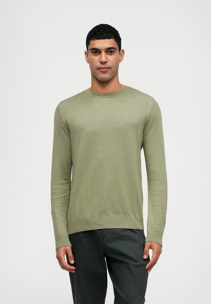 Pull en coton à manches longues couleur vert clair, avec un col rond, des poignets côtelés et une surface texturée ; associé à un pantalon sombre.