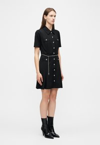 Γυναίκα που φοράει μαύρο φόρεμα με κοντά μανίκια και κουμπιά, με τέσσερις τσέπες και ασημένια ζώνη αλυσίδα, συνδυασμένο με μαύρα μποτάκια αστραγάλου με τακούνι.