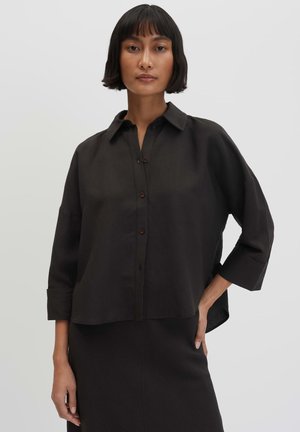 Vrouw met kort donker haar draagt een losse zwarte blouse met knopen en driekwart mouwen en een bijpassende zwarte rok tegen een effen achtergrond.