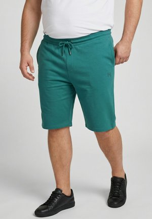 Uomo con pantaloncini verdi con coulisse, sneakers nere e t-shirt bianca, in piedi su uno sfondo chiaro uniforme.