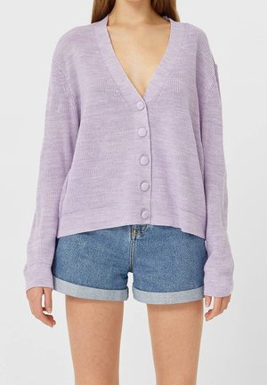 Femme portant un cardigan boutonné violet clair à manches longues et un short en denim bleu retroussé sur un fond uni.