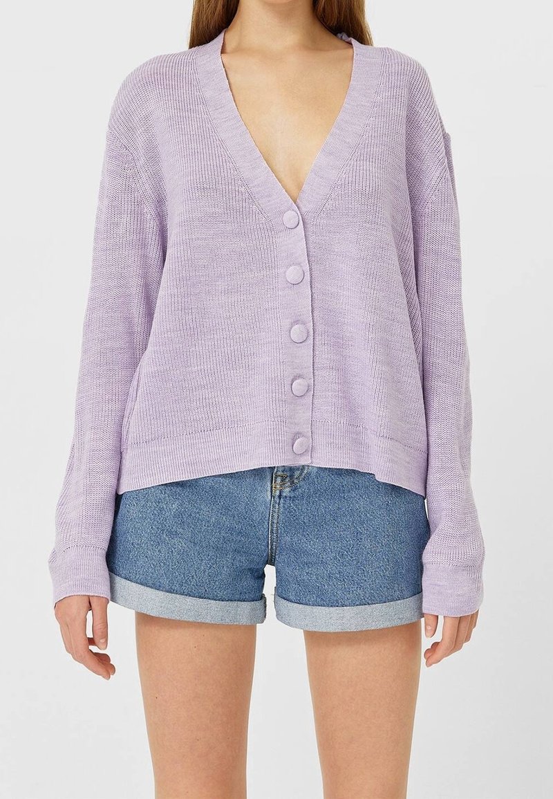 Femme portant un cardigan boutonné violet clair à manches longues et un short en denim bleu retroussé sur un fond uni.